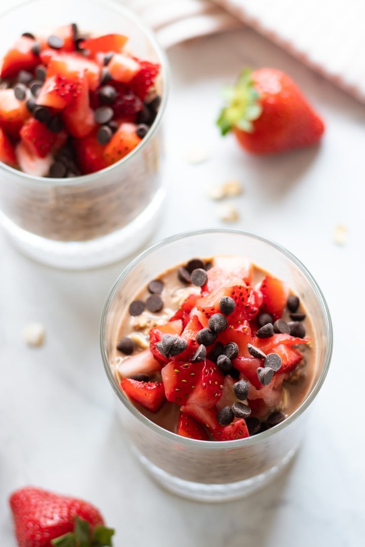 Two glasses filled with low FODMAP chocolate strawberry overnight oats. They are surrounded by a couple of strawberries and in the upper right corner there is light pink striped cloth napkin. 
