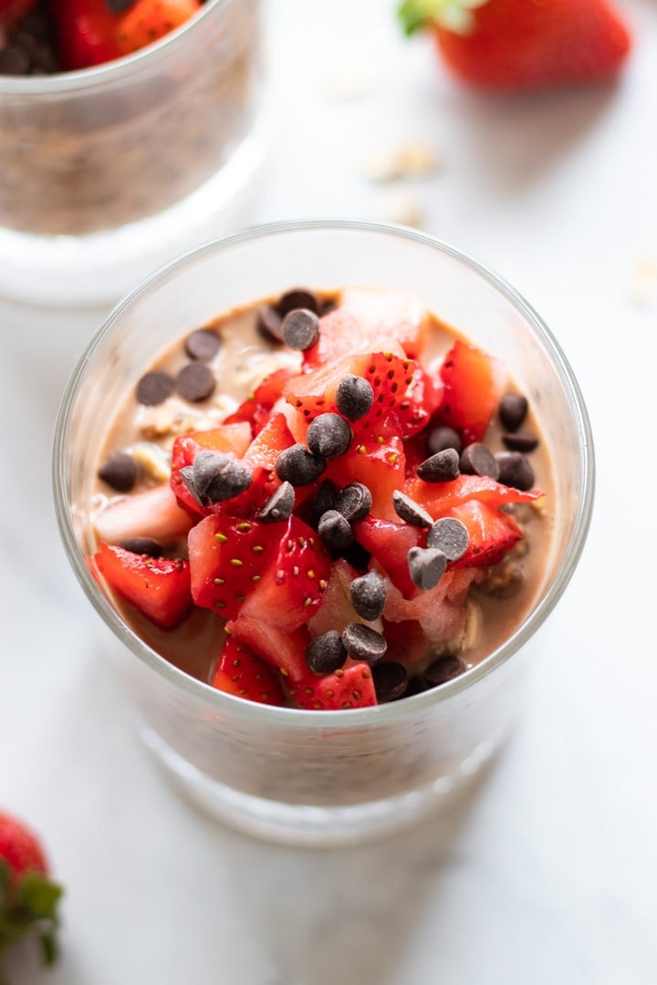 A glass filled with low FODMAP chocolate strawberry overnight oats. It's topped with diced strawberries and mini chocolate chips. In the background there are artistically-blurred out strawberries and rolled oats. 