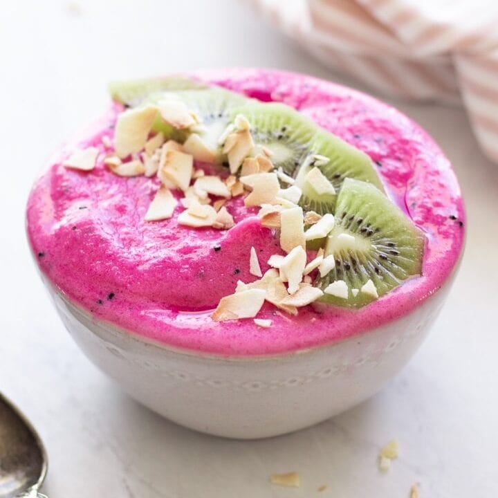 A hot pink low FODMAP dragon fruit smoothie bowl is topped with kiwi slices and coconut flakes in a white bowl. An antique spoon is placed in the foreground and a light pink and white cloth napkin rests in the background.
