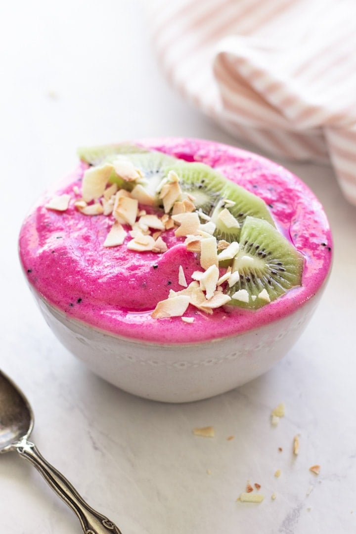 A hot pink low FODMAP dragon fruit smoothie bowl is topped with kiwi slices and coconut flakes in a white bowl. An antique spoon is placed in the foreground and a light pink and white cloth napkin rests in the background.