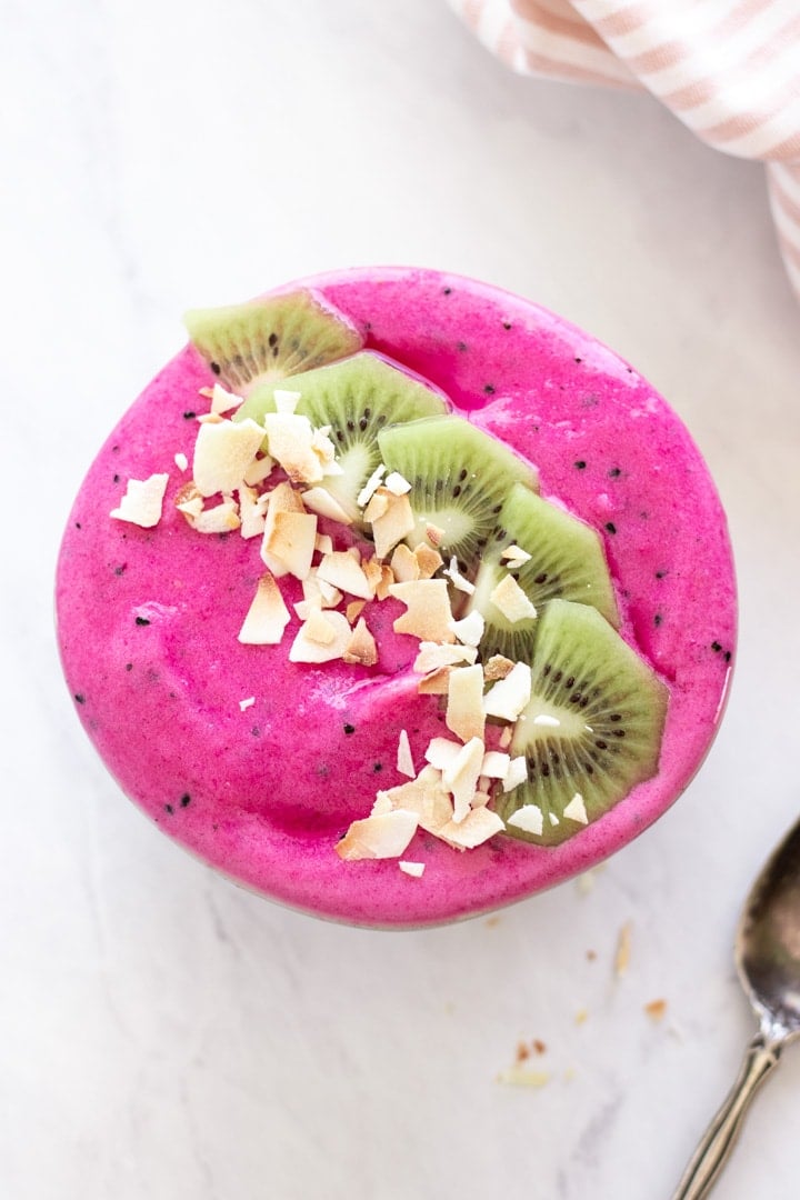 An overhead shot of a hot pink low FODMAP dragon fruit smoothie bowl. Kiwi slices and coconut flakes are artistically placed on top for a pop of color. 