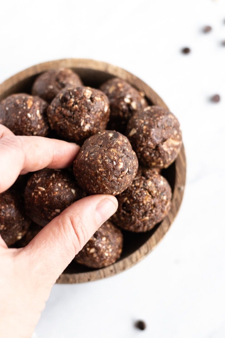 A hand is grabbing a chocolate energy bite out of a full bowl of bites. A couple of chocolate chips are scattered on the marble slab in the background.