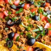A close up of low FODMAP sheet pan nachos or corn chips topped with sliced black olives, diced fresh tomatoes, cilantro, ground turkey, green onion tops, and melty cheddar.