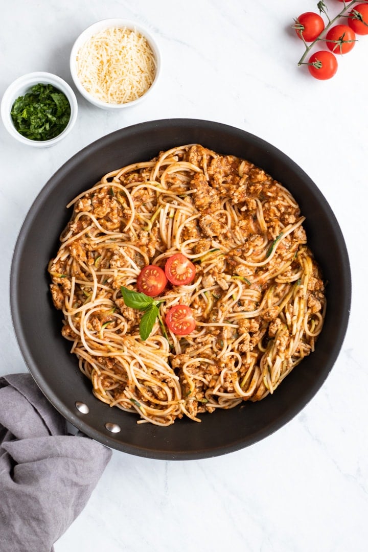 An overhead shot of a black skillet filled with low FODMAP spaghetti and zoodles. The skillet handle is wrapped in a dark gray napkin. A couple of small white dishes filled with parmesan cheese and basil chiffonade frame the skillet. 