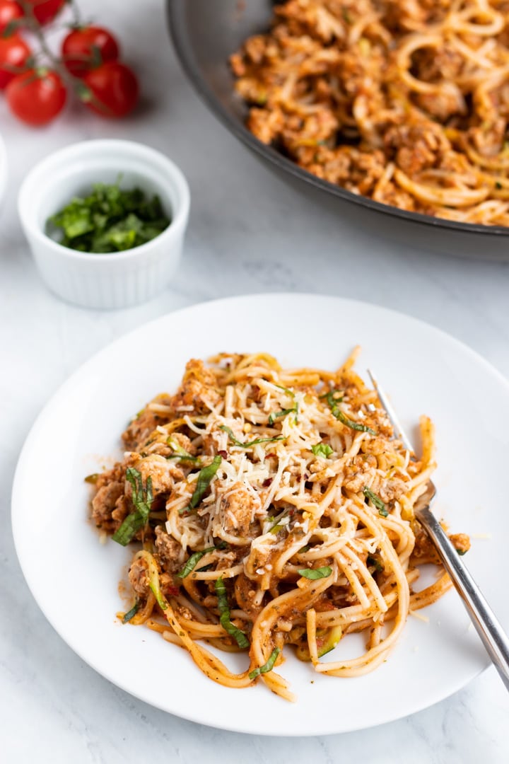 A white plate filled with low FODMAP spaghetti and zoodles sits on a white marble slab. There is a fork set on the plate. A skillet filled with additional pasta, a couple of cherry tomatoes, and a dish filled with shredded basil fill the background.