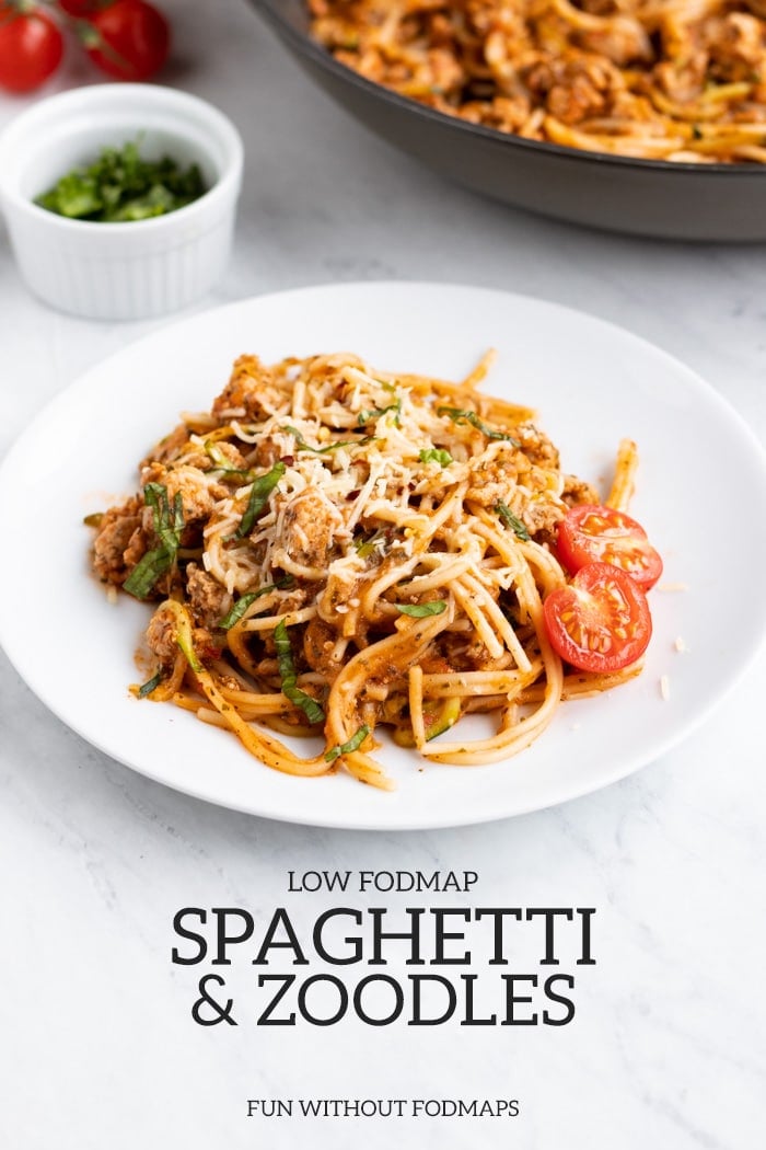 A small white plate is filled with low FODMAP spaghetti and zoodles. In the background there is skillet filled with pasta, a white dish filled with basil chiffonade, and a couple of cherry tomatoes. At the bottom center, dark gray text reads low FODMAP spaghetti and zoodles and below that FUN WITHOUT FODMAPS.