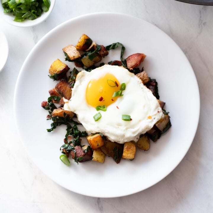 A plate of low FODMAP bacon, chard, and potato hash topped with a sunny-side up egg and sliced green onion tops.