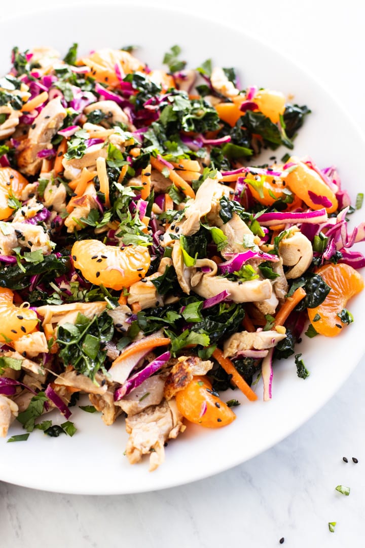 A plate of Chinese chicken salad filled with colorful kale, red cabbage, carrots, mandarin oranges, green onion tops, and chicken.