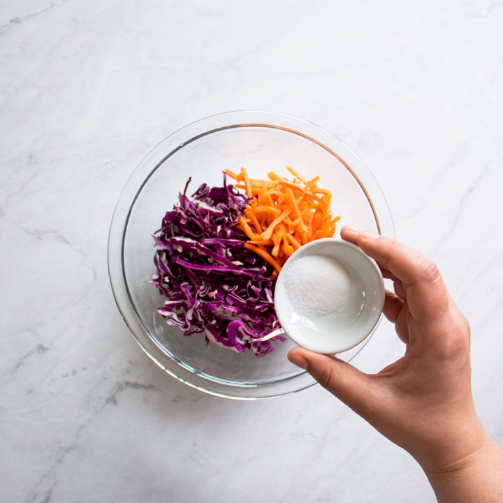 A small bowl of salt is about to be poured into a bowl of shredded red cabbage and carrots.