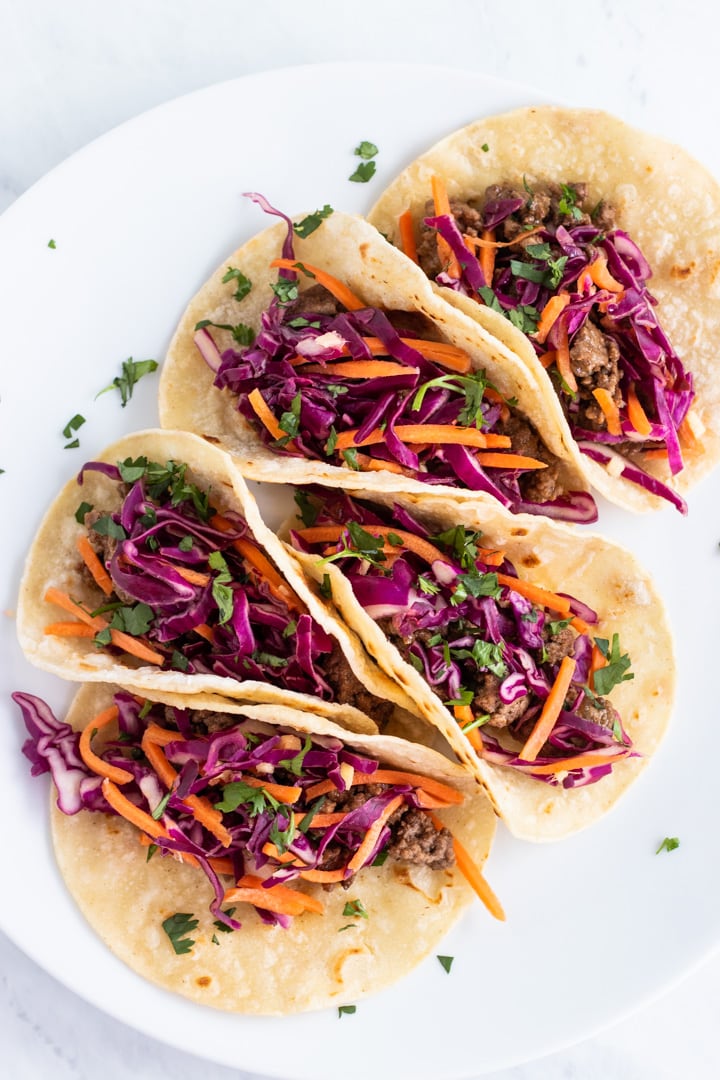 A plate full of five Korean BBQ beef tacos topped with shredded red cabbage, carrot matchsticks, and cilantro.