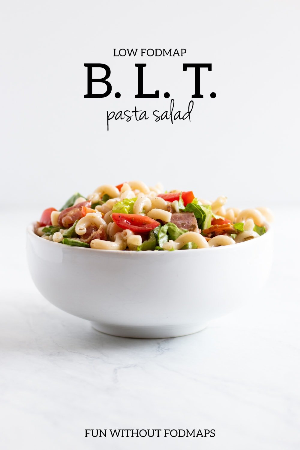 A white bowl filled with a cold pasta and veggie salad sitting on a white marble counter. In the white space above, black text reads "Low FODMAP BLT pasta salad."
