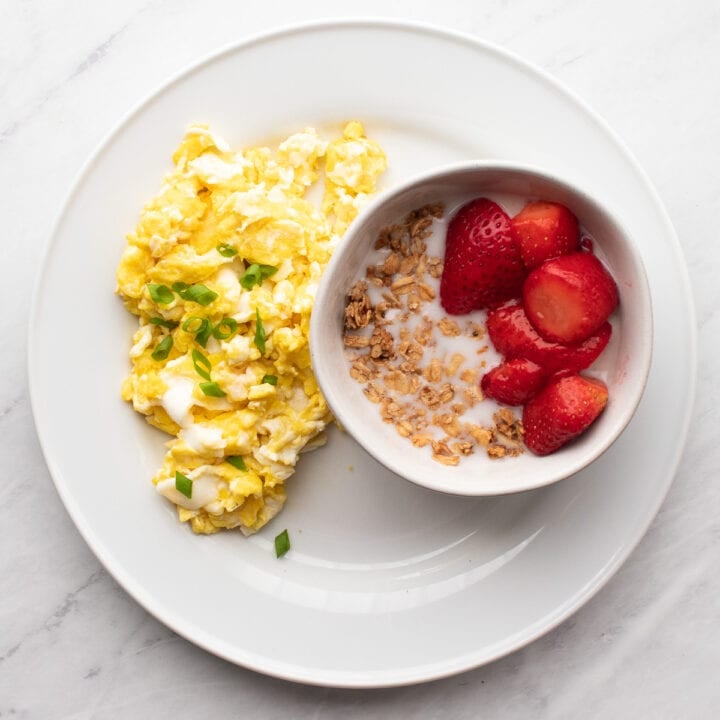 Scrambled eggs with a sprinkle of sliced green onion tops (the low FODMAP part) are sitting on a white plate next to a small bowl filled with maple walnut granola, almond milk, and thawed, frozen strawberries.