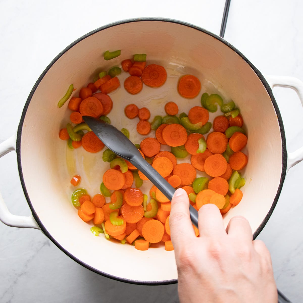 Stirring carrot coins and celery pieces being sautéed in a white Dutch oven pot.