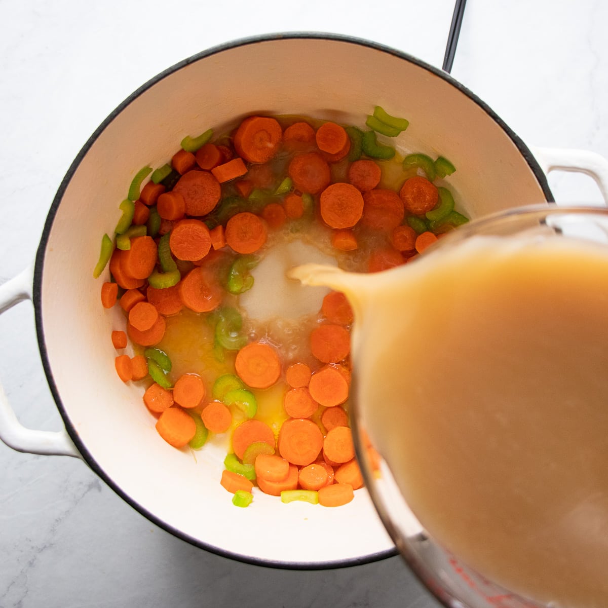 Pouring low FODMAP chicken broth into a Dutch oven containing sautéed carrots and celery pieces.