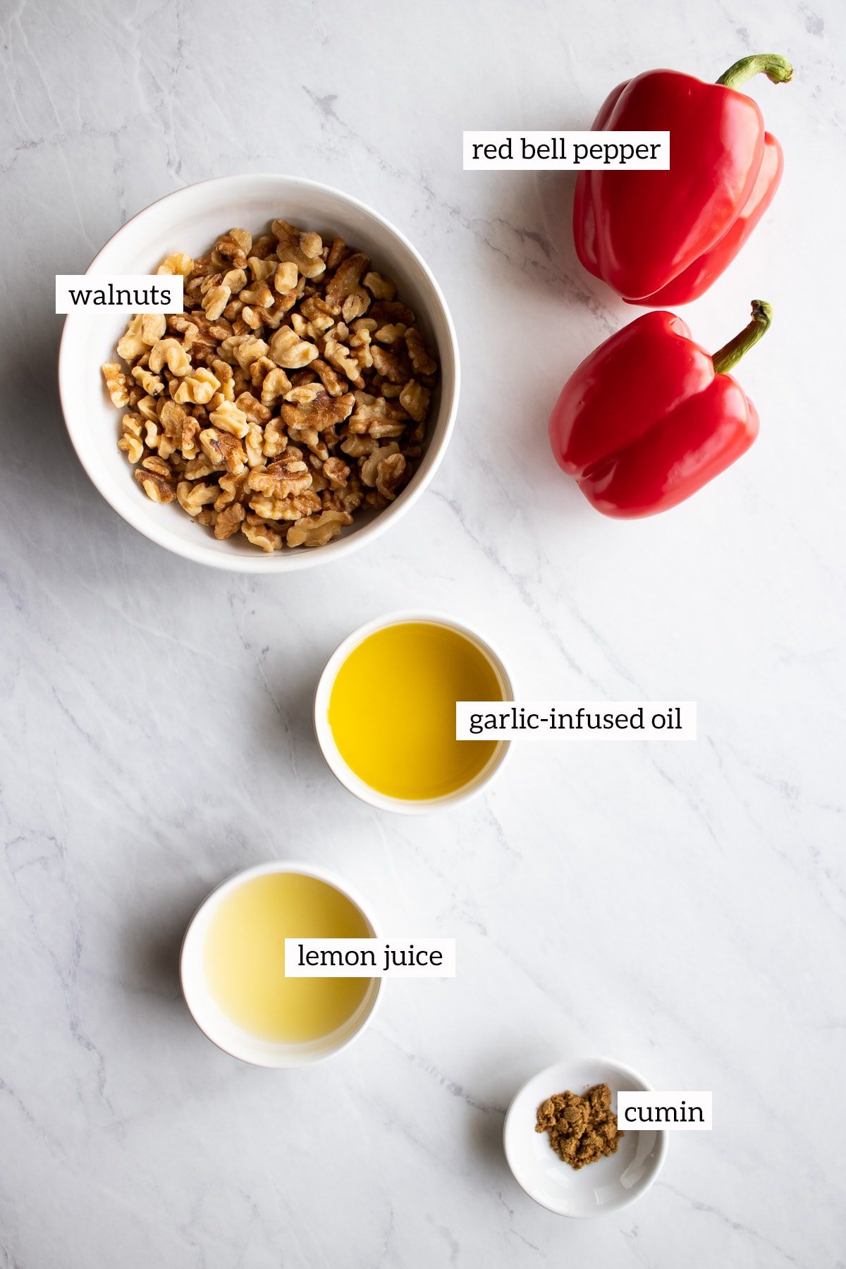 Ingredients needed for this low FODMAP red pepper and walnut dip are measured out into white bowls on a white marble slab.