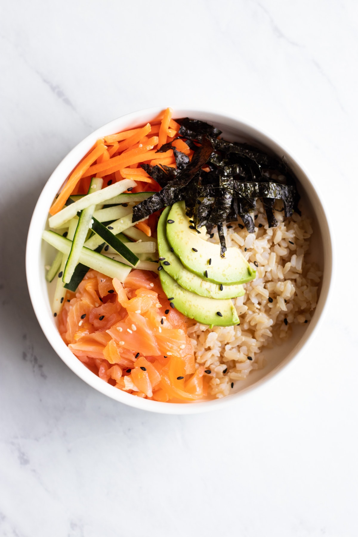 A single white bowl filled with a base of brown rice, small handfuls of smoked salmon, cucumber matchsticks, carrot matchsticks, and nori (seaweed) strips are arranged on the left side of the bowl. Several avocado slices are in the center and there's a sprinkle of sesame seeds.