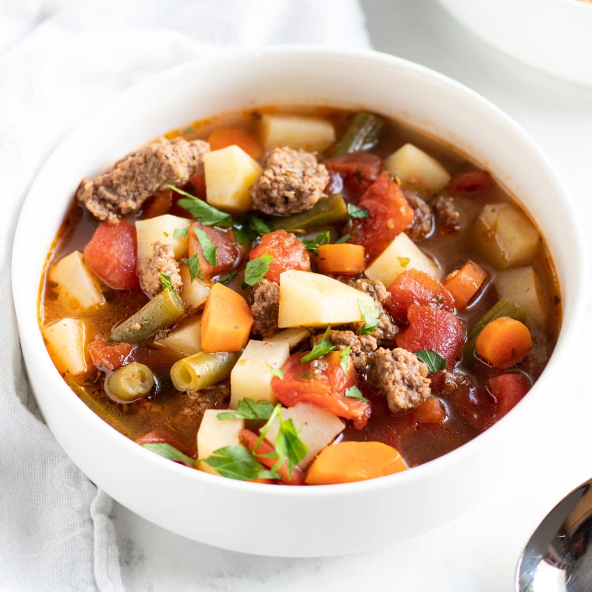 A bowl of soup made with ground beef, potatoes, carrots, tomatoes, and green beans.