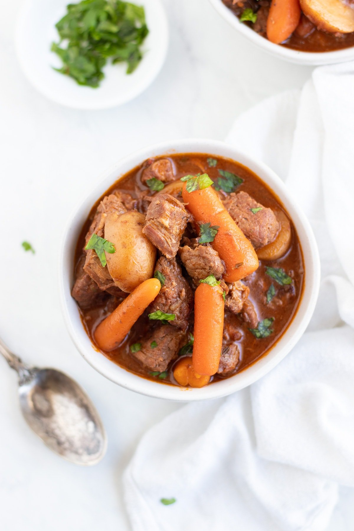 A small bowl of beef stew surrounded by a spoon and pinch bowl filled with chopped fresh parsley.