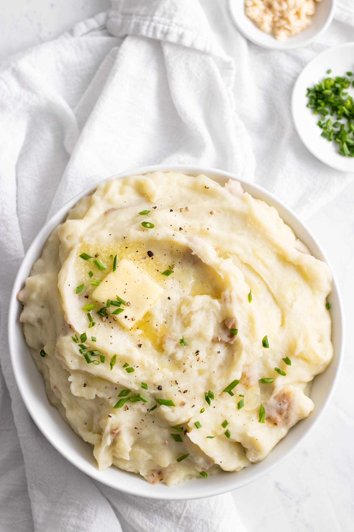 Looking down at a bowl of mashed potatoes topped with snipped fresh chives, a dab of melting butter, and freshly-cracked black pepper. The bowl sits on a white linen napkin and is surrounded by small bowls of snipped chives and grated horseradish.