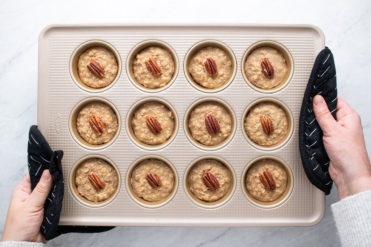 Hands protected by pot holders are holding the muffin tin filled with the baked oatmeal cups.