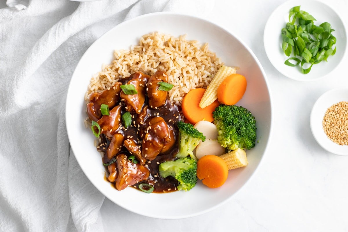 A bowl of sesame chicken served over brown rice with a side of steamed broccoli, carrots, baby corn, and water chestnuts.