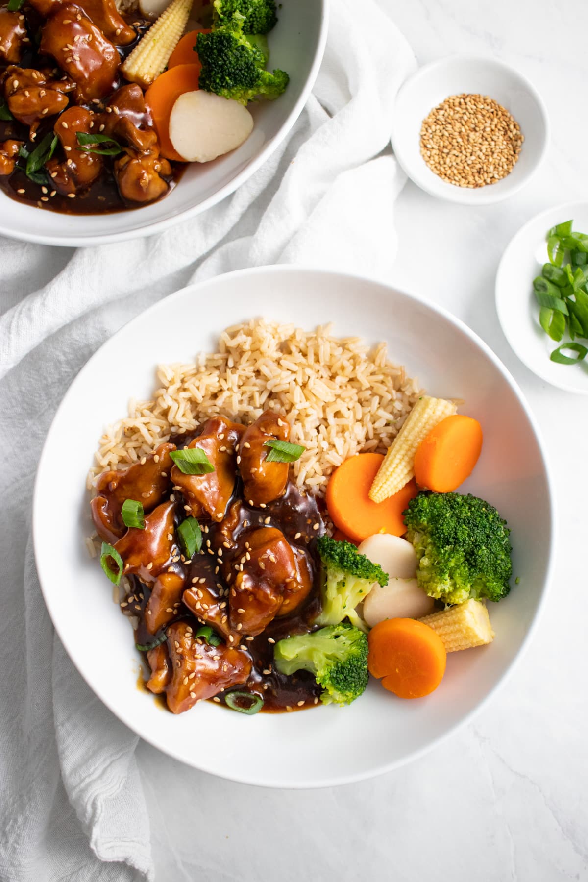 Looking down at a bowl of sesame chicken served with brown rice and steamed veggies. A second bowl of chicken, as well as small bowls of toasted sesame seeds and sliced green onion tops sit above it.