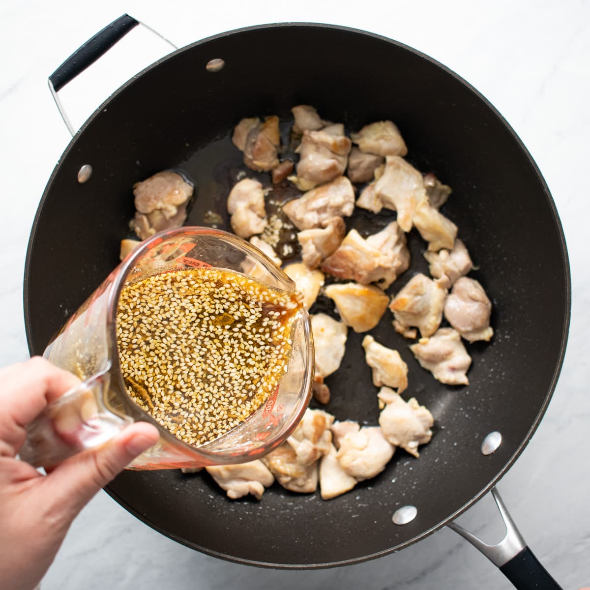Pouring sesame sauce into a skillet with cooked chicken pieces.