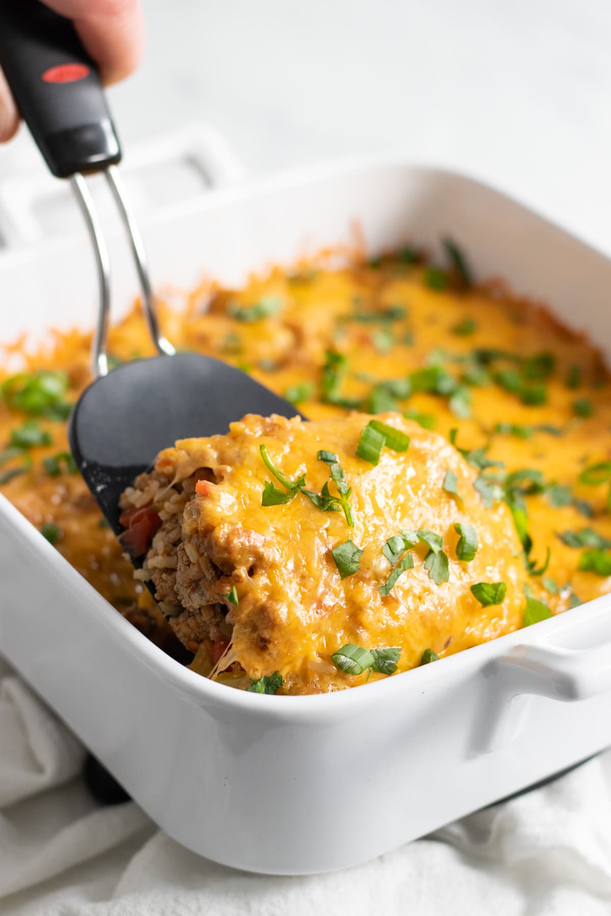 A black spatula lifting a serving of cheesy casserole made with ground turkey, brown rice, chiles, and tomatoes out of a square white baking dish.
