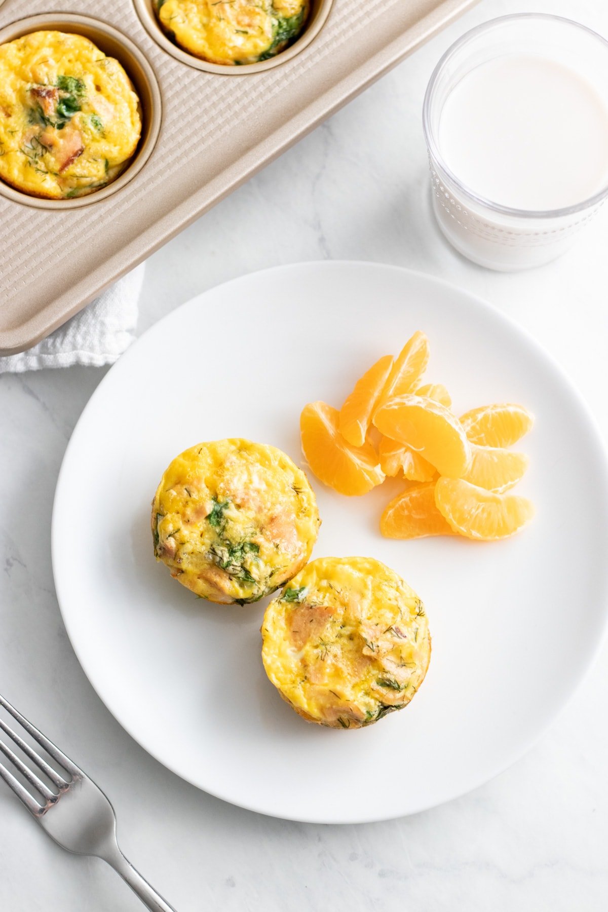 Two smoked salmon and dill egg cups  on a small plate with orange segments. A glass of lactose-free milk and a muffin tin with more egg cups sit nearby.