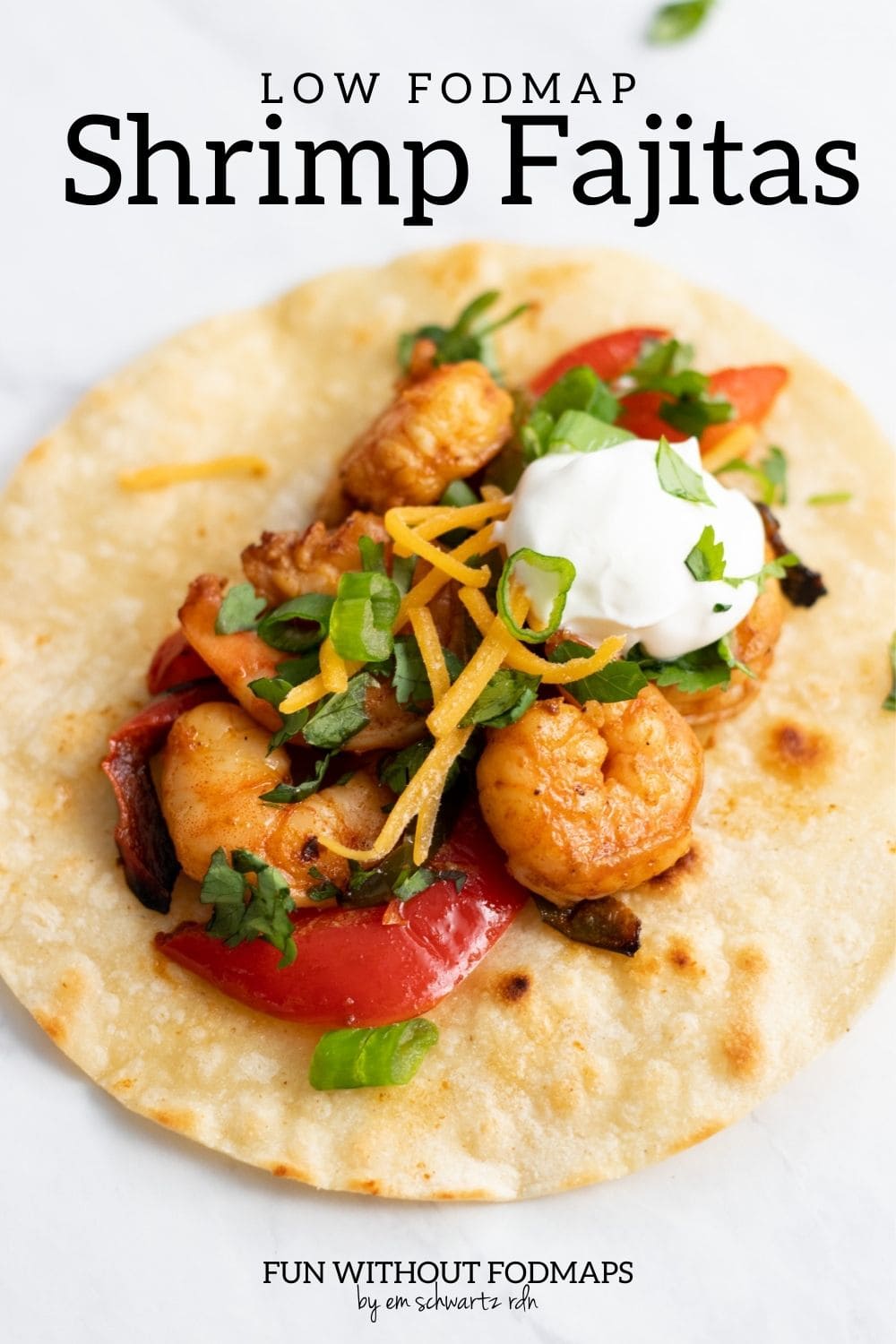 A corn tortilla topped with sautéed shrimp and bell peppers, shredded cheddar cheese, sour cream, cilantro, and sliced green onion tops. In the white space above, a black text overlay reads "Low FODMAP Shrimp Fajitas."