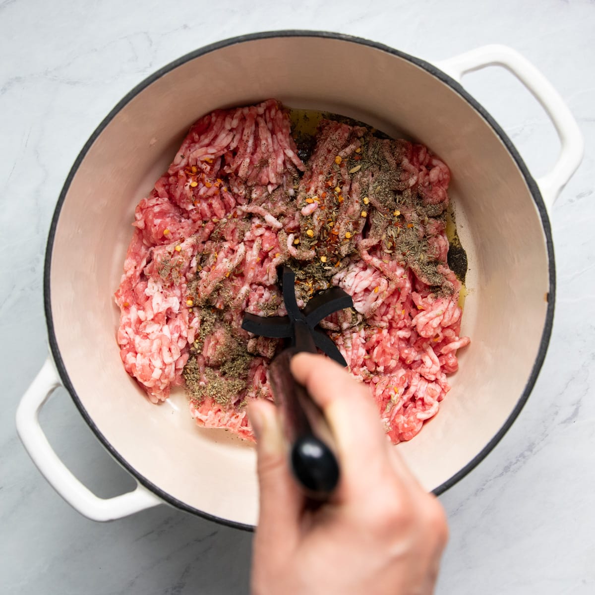 A hand is breaking up ground pork in a Dutch oven with a meat masher
