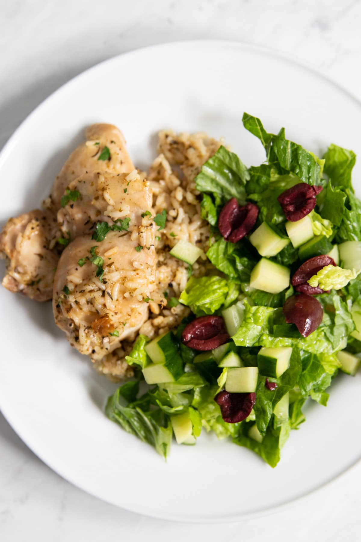 A white plate with a serving of low FODMAP lemon chicken and rice, as well as a side salad of chopped romaine, halved olives, and diced cucumber. 