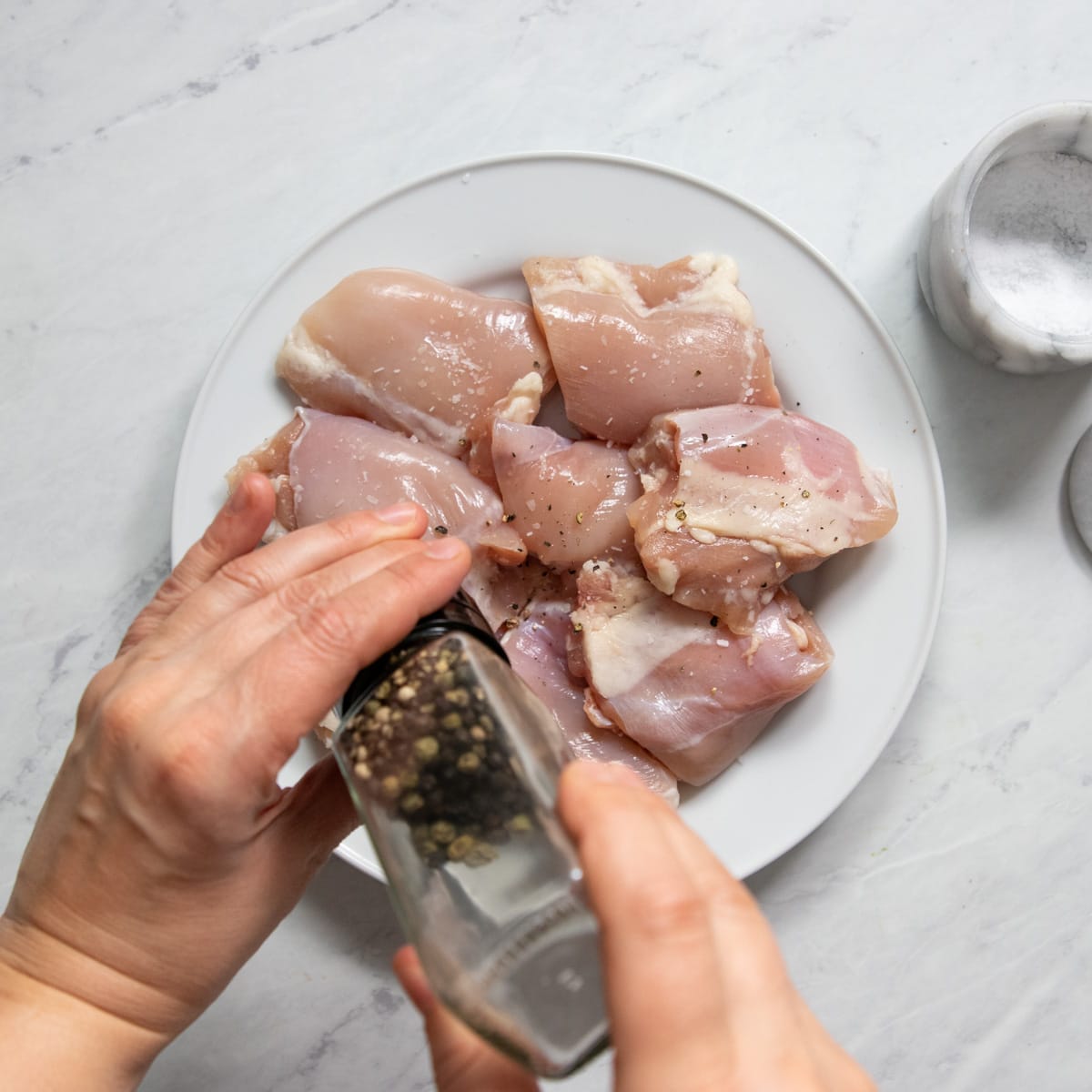 Hands are grinding a glass black pepper mill over a plate of boneless, skinless chicken thighs.