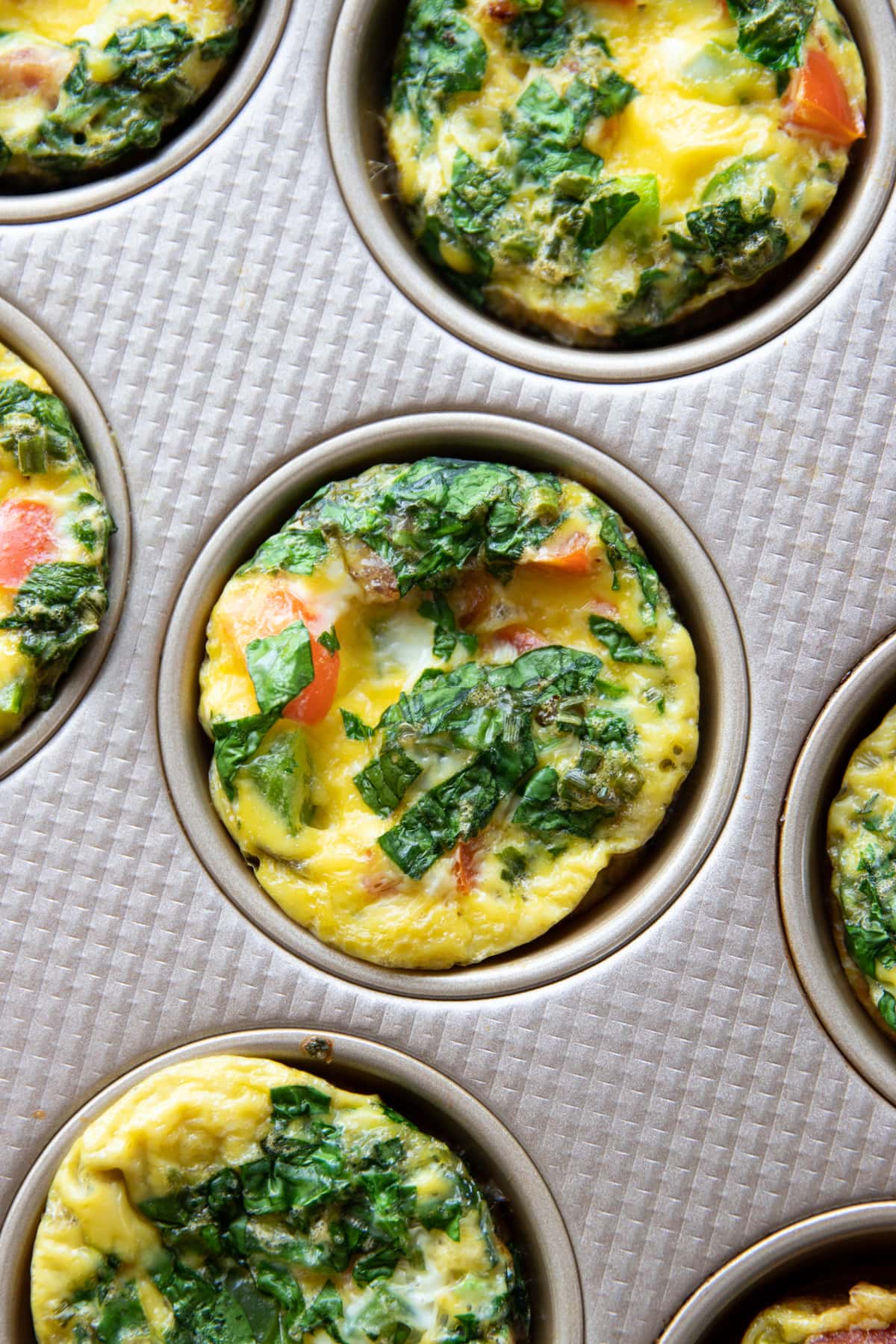 Zoomed into a baked egg muffin in a muffin tin. It's made with chopped spinach, dried chives, and red and green bell pepper.