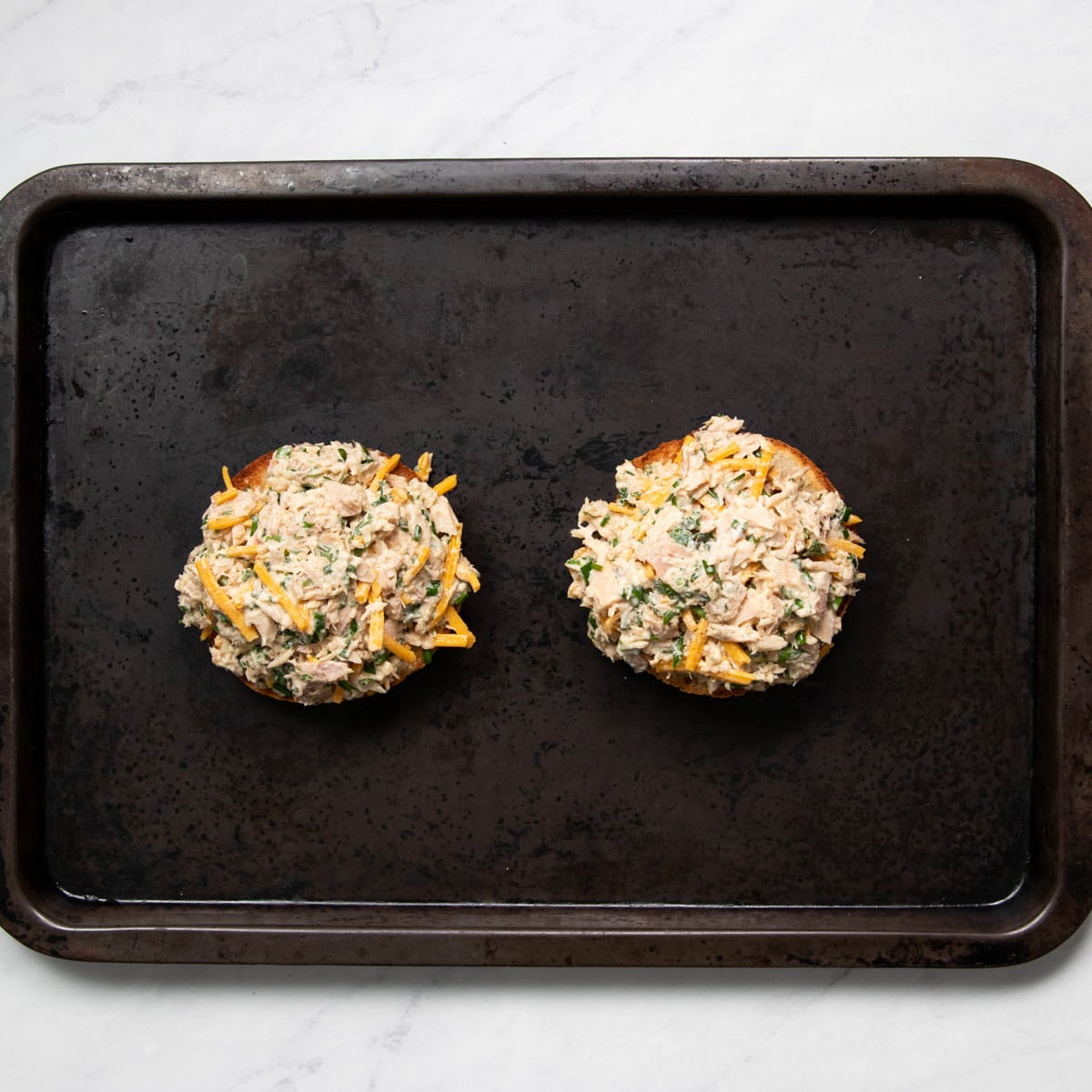 Prepared tuna salad is divided onto two toasted English muffin halves on a baking sheet
