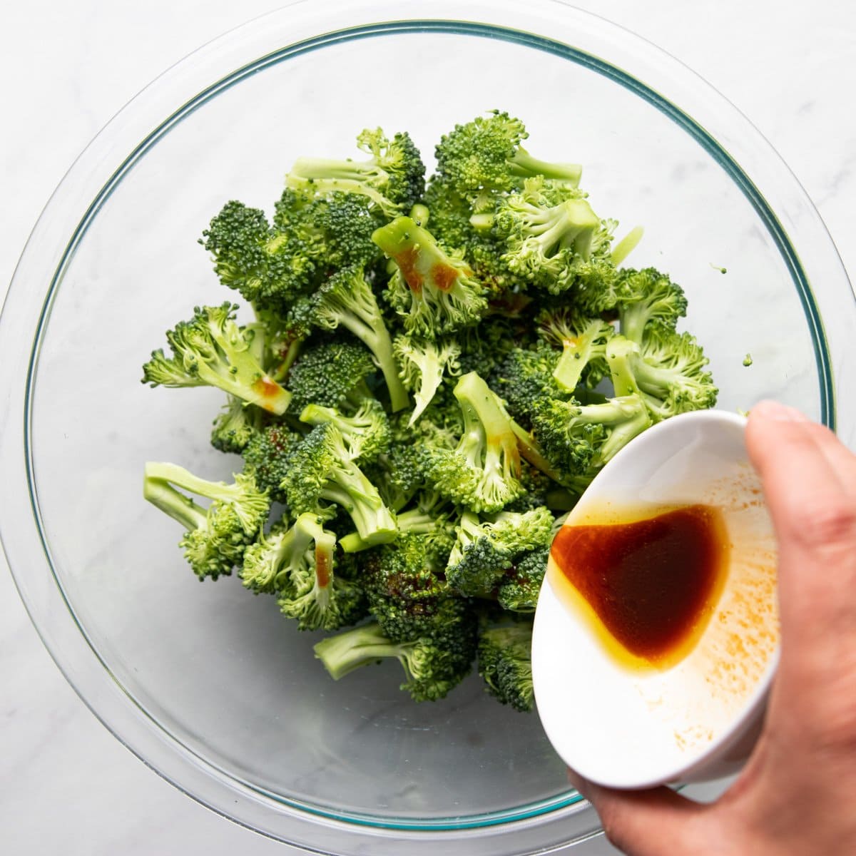 Pouring sauce onto broccoli florets in a large glass mixing bowl. 