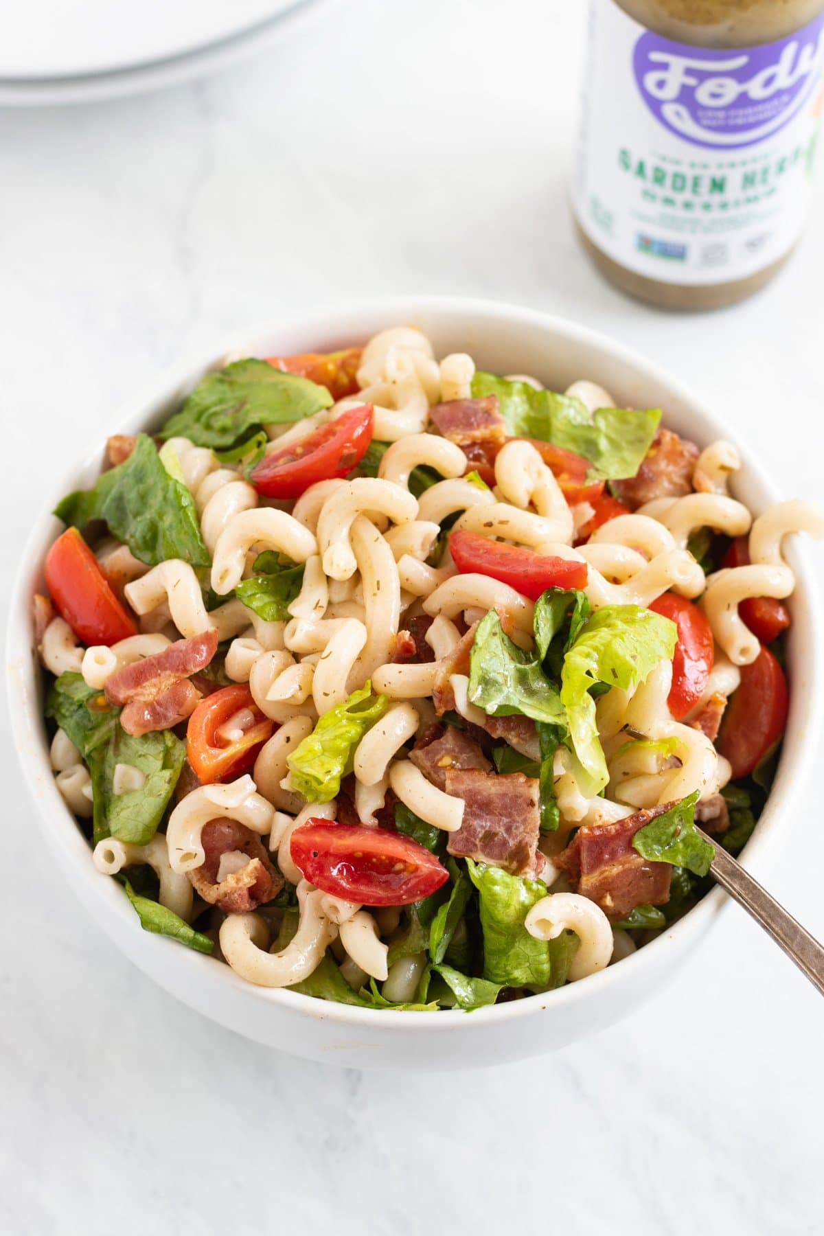 A bowl of blt pasta salad with a bottle of Fody Foods Garden Herb Dressing in the background.