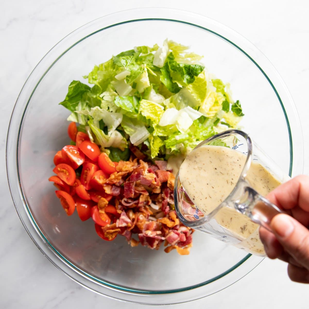 Pouring Fody's Herb Dressing into a mixing bowl filled with lettuce, cooked pasta, tomatoes, and crumbled bacon.