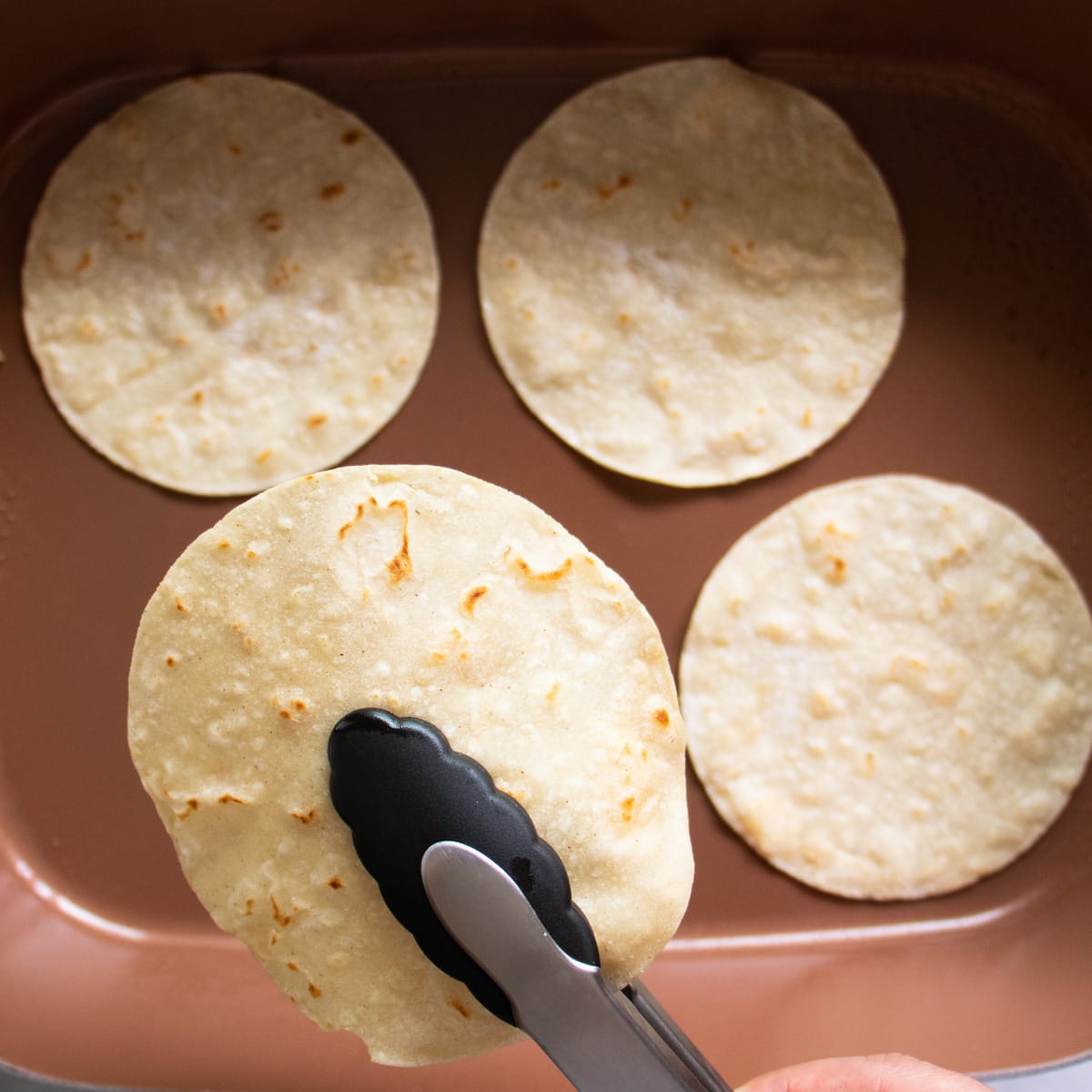 Flipping corn tortillas on an electric griddle with tongs.