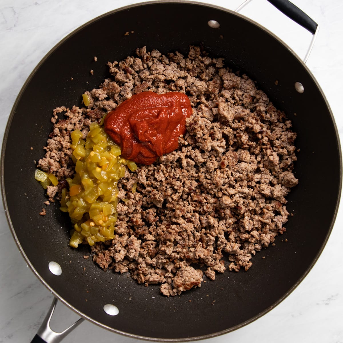 A skillet filled with browned ground beef, diced green chiles, and a homemade tomato sauce ready to be mixed. 