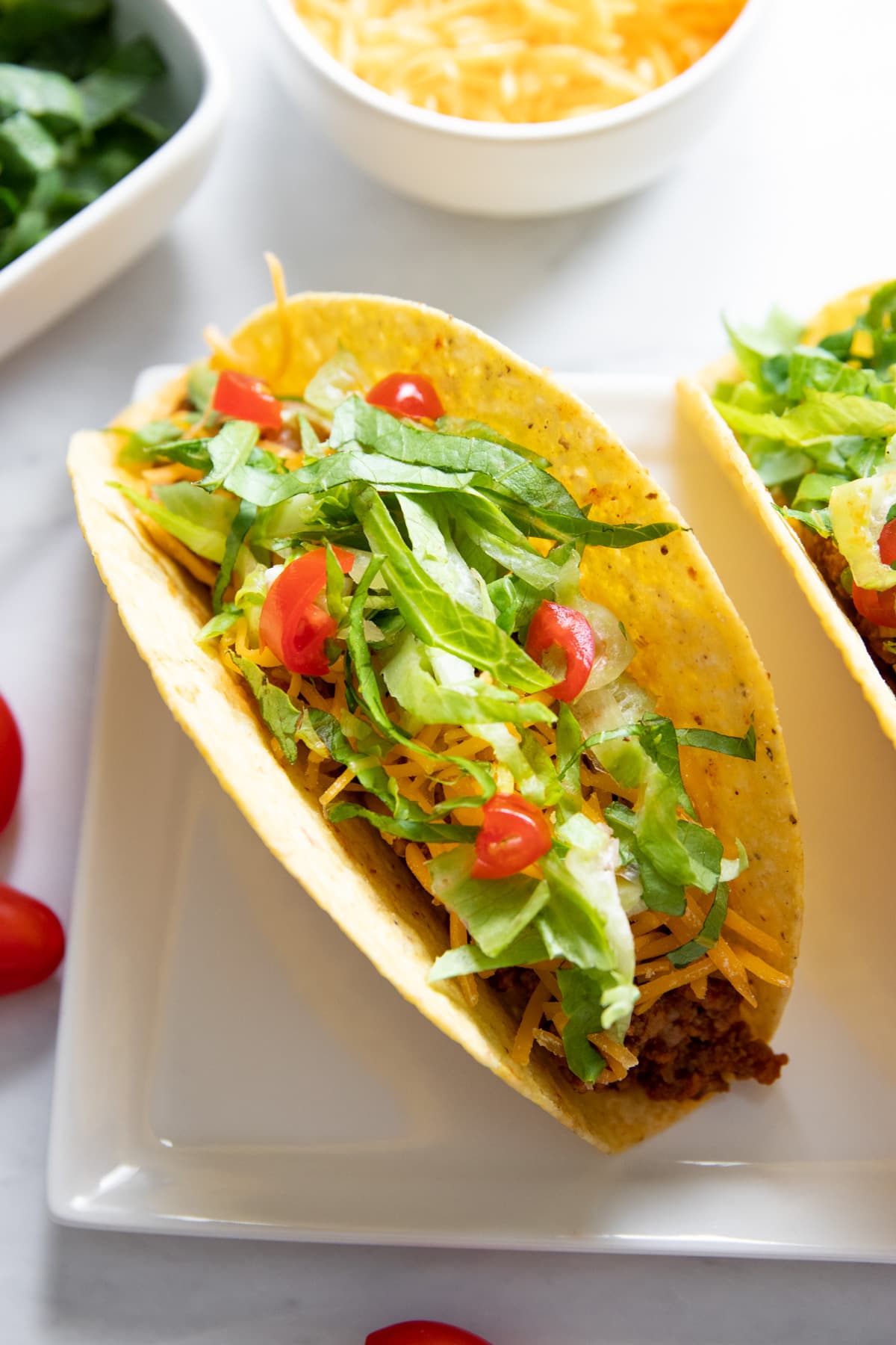 A hard shell taco sits on a rectangular plate. It's surrounded by  small dishes of shredded lettuce and shredded cheddar cheese, as well as a couple of whole cherry tomatoes for decoration.
