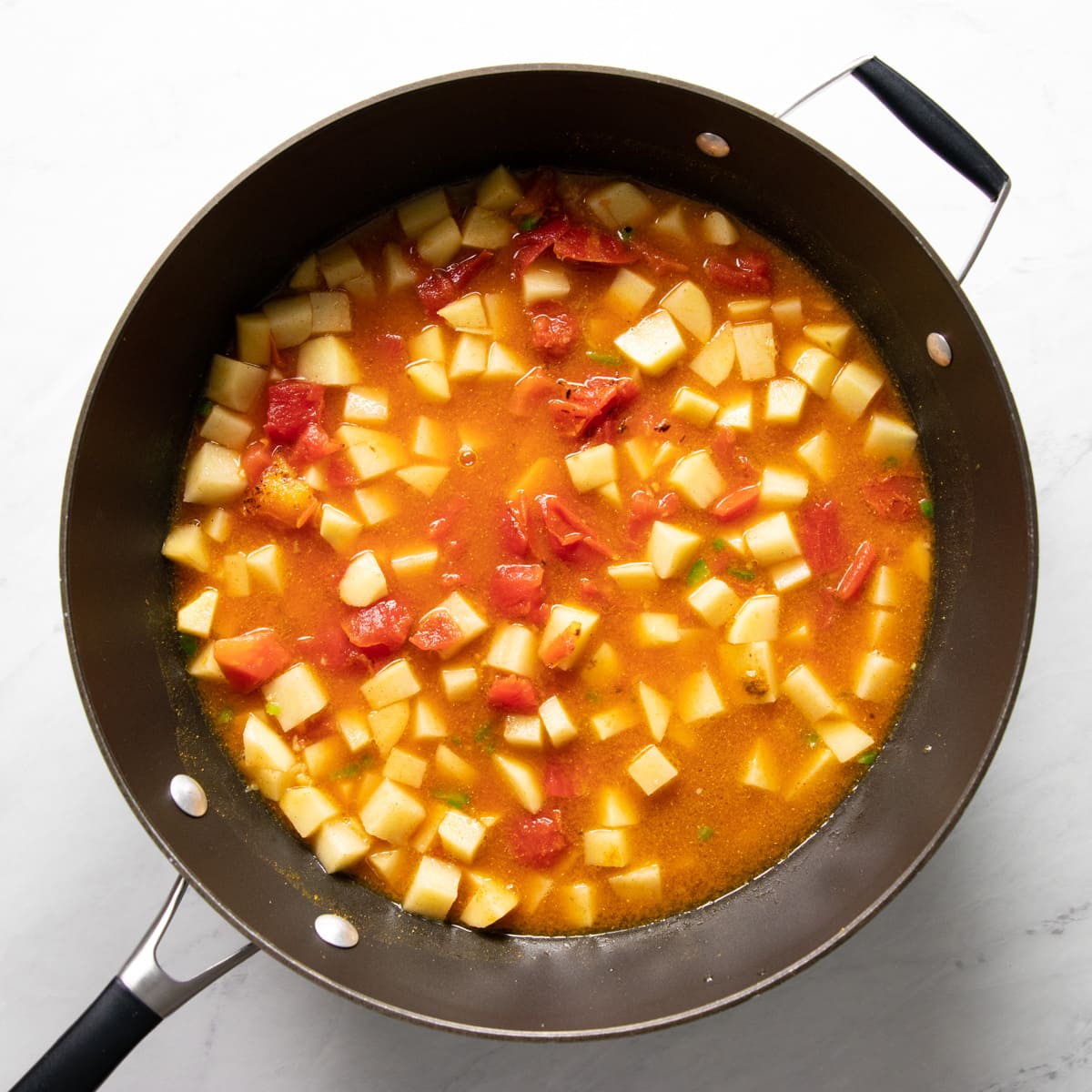 A skillet filled with canned tomatoes, uncooked diced potatoes, and a vegetarian broth ready to simmer. 
