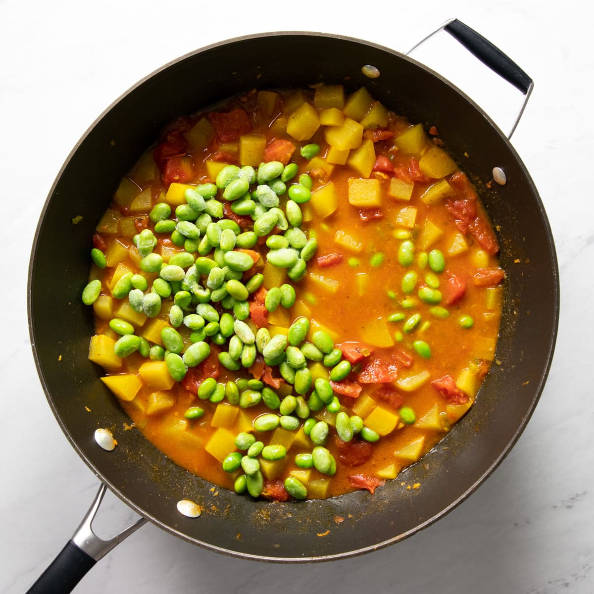 Frozen shelled edamame have just been added to a skillet filled with a cooked curry containing diced potatoes and canned tomatoes. 