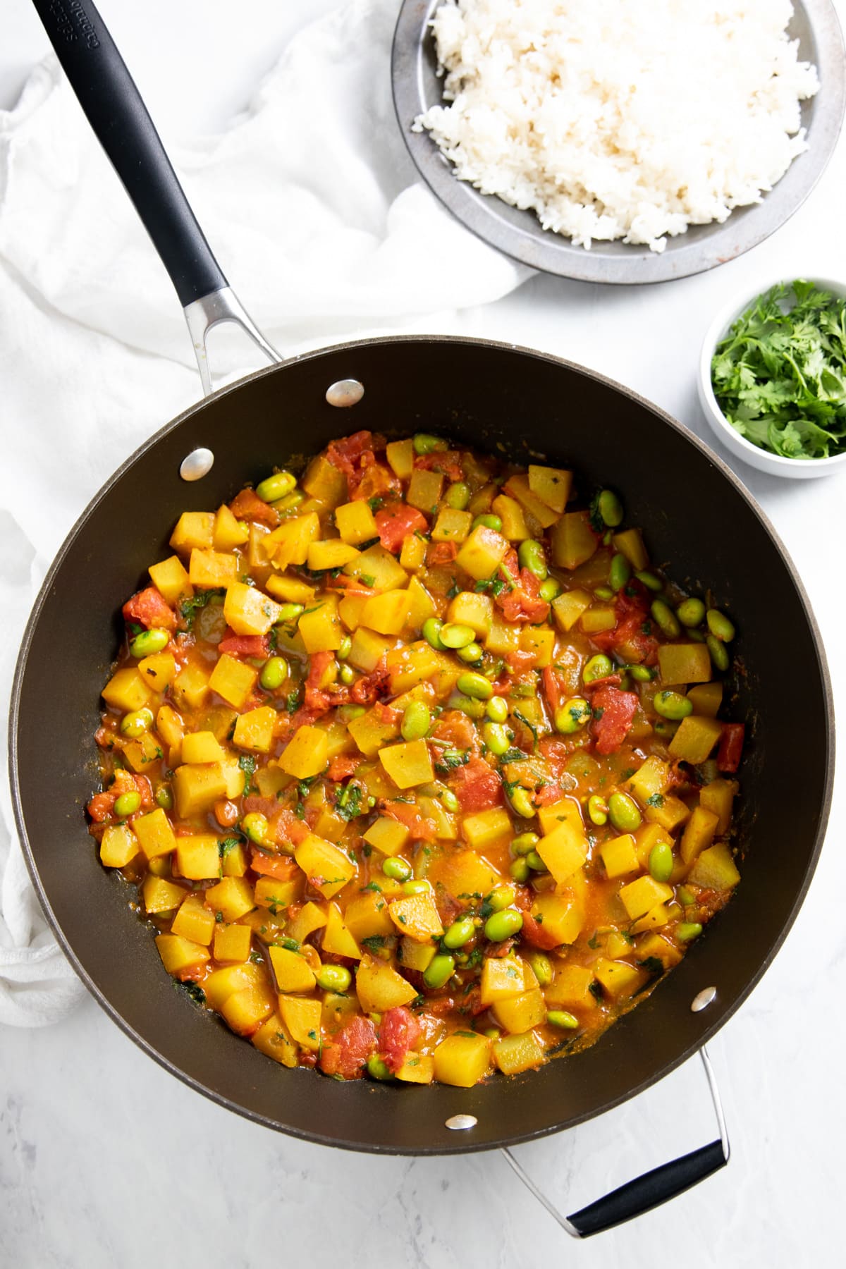 A skillet is filled with a curry made with diced Yukon gold potatoes, canned fire-roasted tomatoes, and shelled edamame. Smaller dishes of cooked white rice and chopped cilantro sit in the upper right corner. 