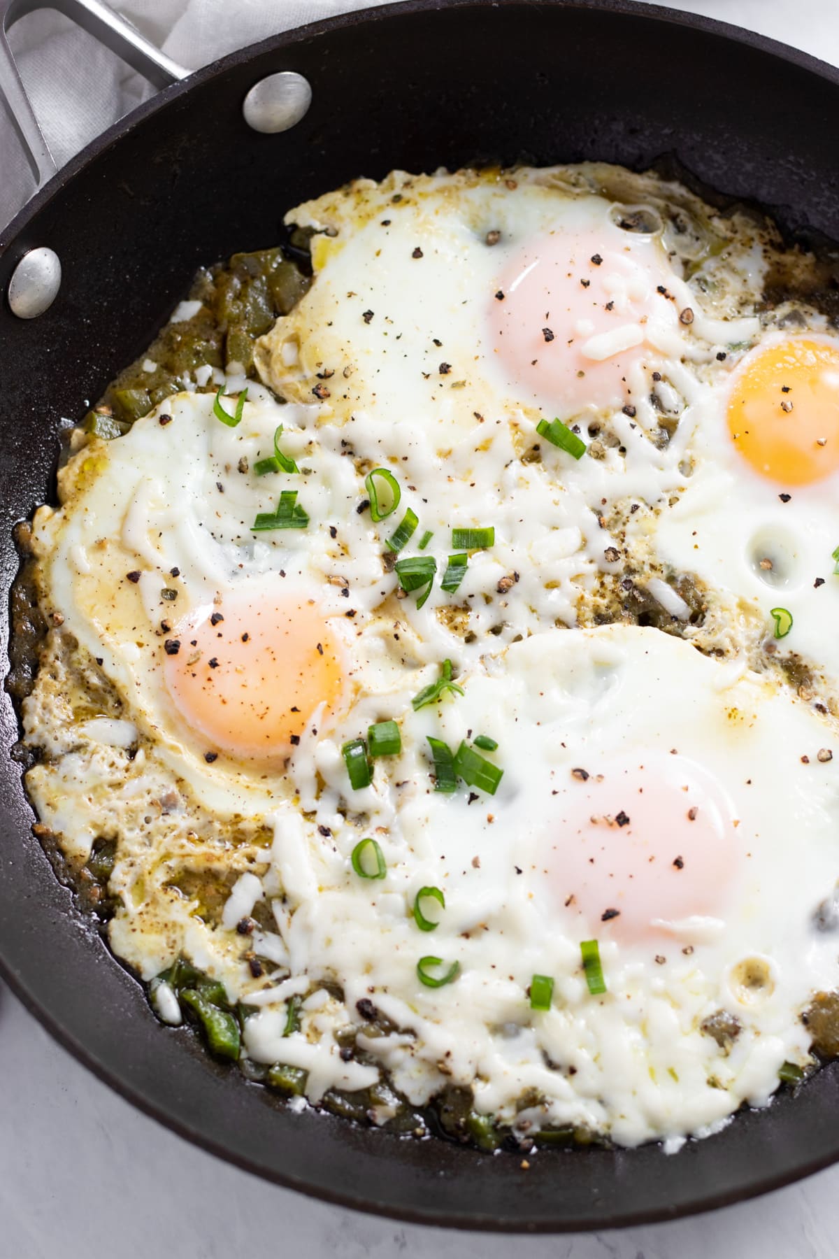 Zoomed into a skillet filled with steam-cooked eggs in salsa verde. The eggs are garnished with sliced green onion tops, melty Oaxaca cheese, and black pepper.