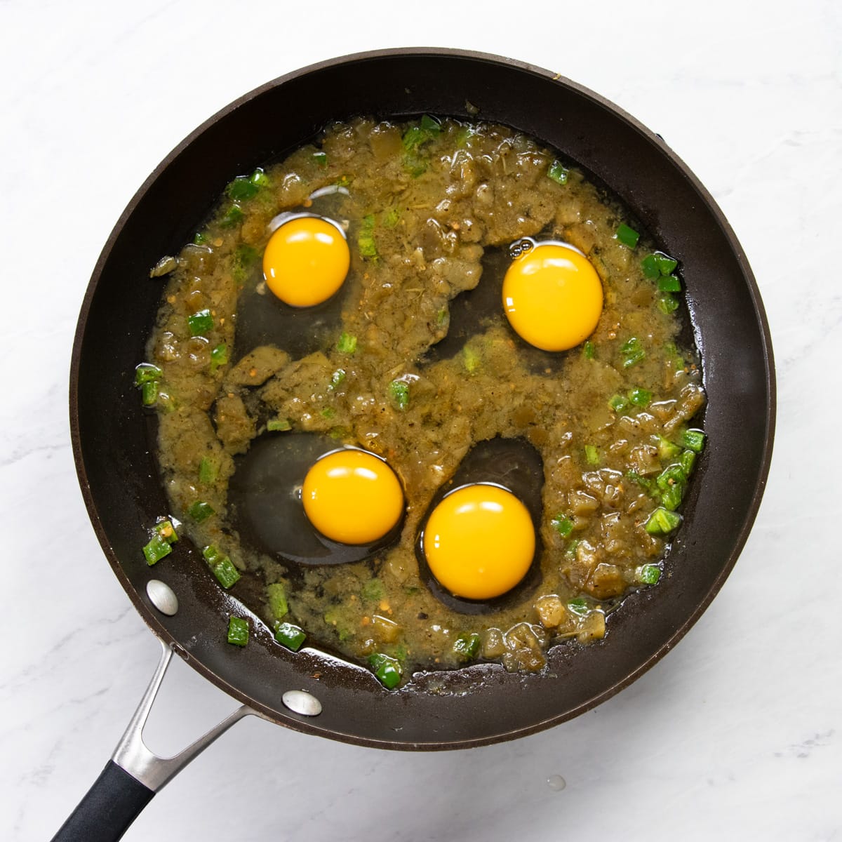 A skillet with four raw eggs cracked into salsa verde ready to be steam-cooked.