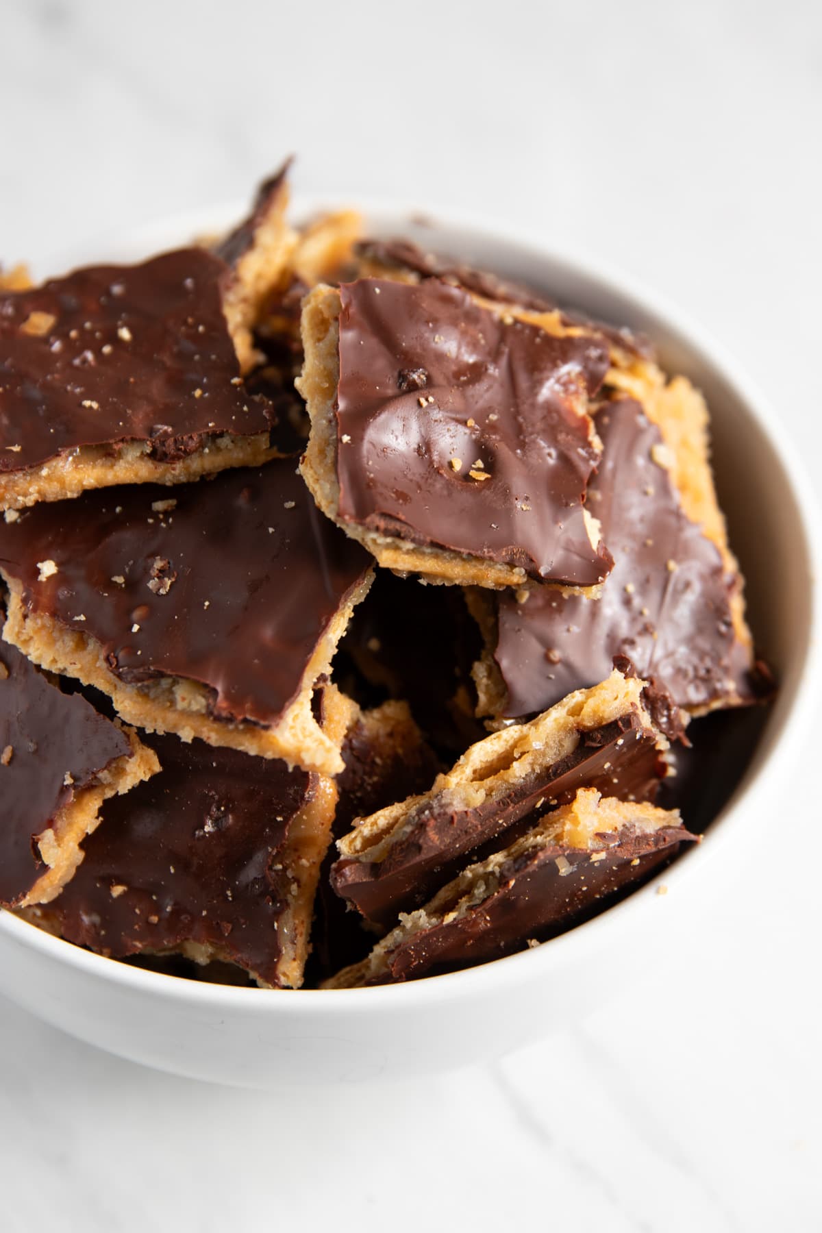A white bowl containing pieces of cracker toffee each topped with a thin layer of dark chocolate.