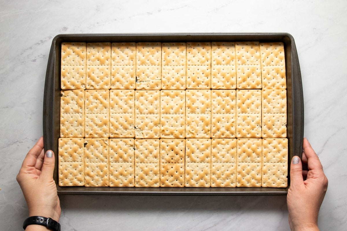 Hands holding a sheet pan filled with a single layer of Schar Gluten Free Table Crackers.