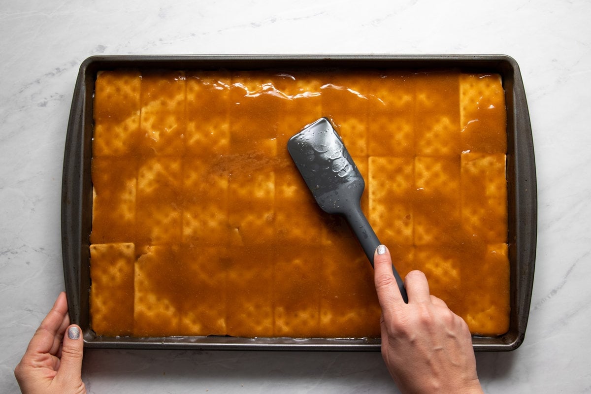 Spreading homemade melted toffee with a silicone spatula on top of a sheet pan lined with an even layer of table crackers.