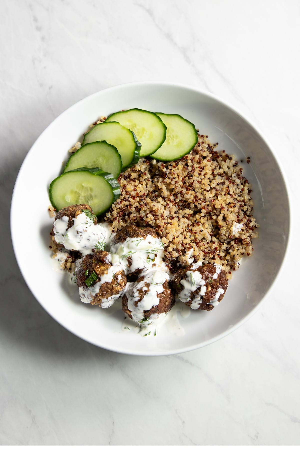 A shallow bowl filled with cooked quinoa, low FODMAP Greek meatballs topped with a creamy dill sauce, and a few cucumber slices on the side.